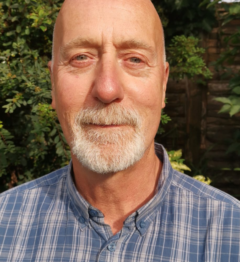 Photo of Richard Spong wearing a blue and white checked shirt, stood in front of greenery.